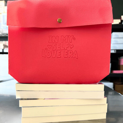 In my self Love Era is printed on a red bag sitting atop of five books with their pages facing out