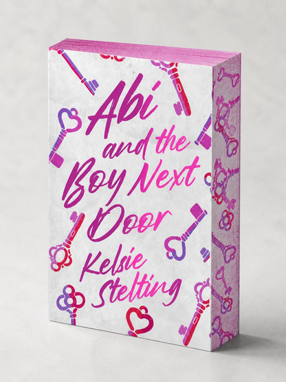 A high-resolution 3D mockup of the paperback for Abi and the Boy Next Door by Kelsie Stelting.

Cover Design: Features a white textured background with "Abi and the Boy Next Door" written in a vibrant pink and purple gradient script. The cover is decorated with various illustrated keys in pink and purple hues.

Sprayed Edges: The book is angled to show the top and side edges, which are sprayed with a bright pink color and a repeating purple key pattern, matching the cover's aesthetic.